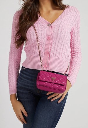 Kobieta ubrana w różowy sweter na druty z warkoczowym splotem i ciemne dżinsy, niosąca małą pikowaną różową torebkę Guess na ramię z łańcuszkowym paskiem.