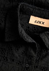 Μαύρο υφασμάτινο πουκάμισο με περίπλοκα κεντημένα μοτίβα, μπροστινό κούμπωμα με κουμπιά και μπεζ ετικέτα με την επιγραφή "JJXX" κοντά στον γιακά.
