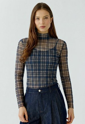 Vrouw met lang bruin haar die een doorschijnende geruite longsleeve draagt over een donkerder geruite tanktop, gecombineerd met hooggetailleerde donkerblauwe spijkerbroek.