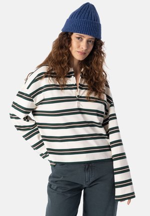 Frau mit blauer Strickmütze, langärmligem weißen Shirt mit grünen Streifen und dunkler Hose steht vor einem schlichten, hellen Hintergrund.