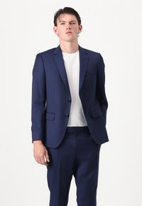Traje azul marino con una americana de un solo botón, solapas en pico y dos botones. Combinado con una camiseta blanca de cuello redondo y pantalones a juego.