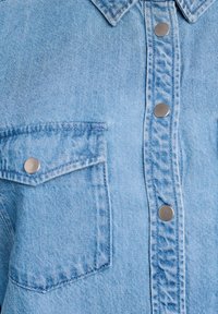 Svetlomodrá džínsová košeľa vpredu s golierom, viditeľná ľavá náprsní kapsa so strieborným patentným gombíkom a tromi striebornými patentnými gombíkmi na prednej légovej časti.