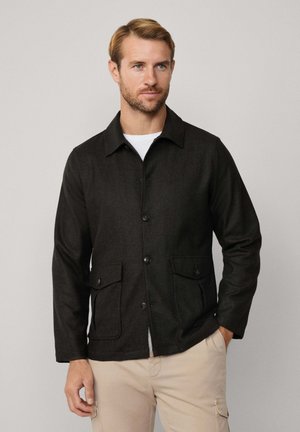 Dunkel gefärbte, gewobene Stoffjacke mit Kragen, vorderer Knopfverschluss und zwei großen, geknöpften Taschen; hat eine taillierte Passform und lange Ärmel.