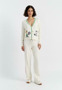 Crèmekleurige cardigan met groene rand en knoopsluiting, met een cartoonafbeelding van personages die tennis spelen. Gecombineerd met bijpassende crèmekleurige broek.