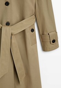 Trench-coat beige avec des boutons noirs, une ceinture nouée à la taille, une poche sur le côté et une sangle de manchette avec un bouton sur la manche.