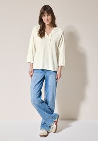 Haut léger couleur crème avec un col en V et des cordons, associé à un pantalon en jean large bleu clair et des baskets blanches avec des accents rouges.