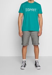 Tyrkysové tričko s krátkým rukávem s logem "ESPRIT", doplněné o srolované šedé šortky a černé vysoké tenisky. Jednoduchý, ležérní outfit.