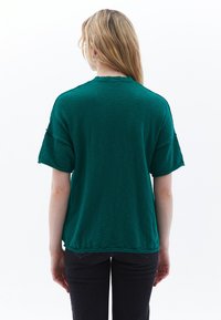 Groene T-shirt met korte mouwen en een ontspannen pasvorm, met een gebogen zoom en subtiele textuur, gedragen met donkere jeans. Achteraanzicht.