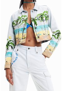 Žena má na sobě zkrácenou džínovou bundu s barevným potiskem pláže, modrý vzorovaný bikiny top, bílé kalhoty a zelený choker náhrdelník.