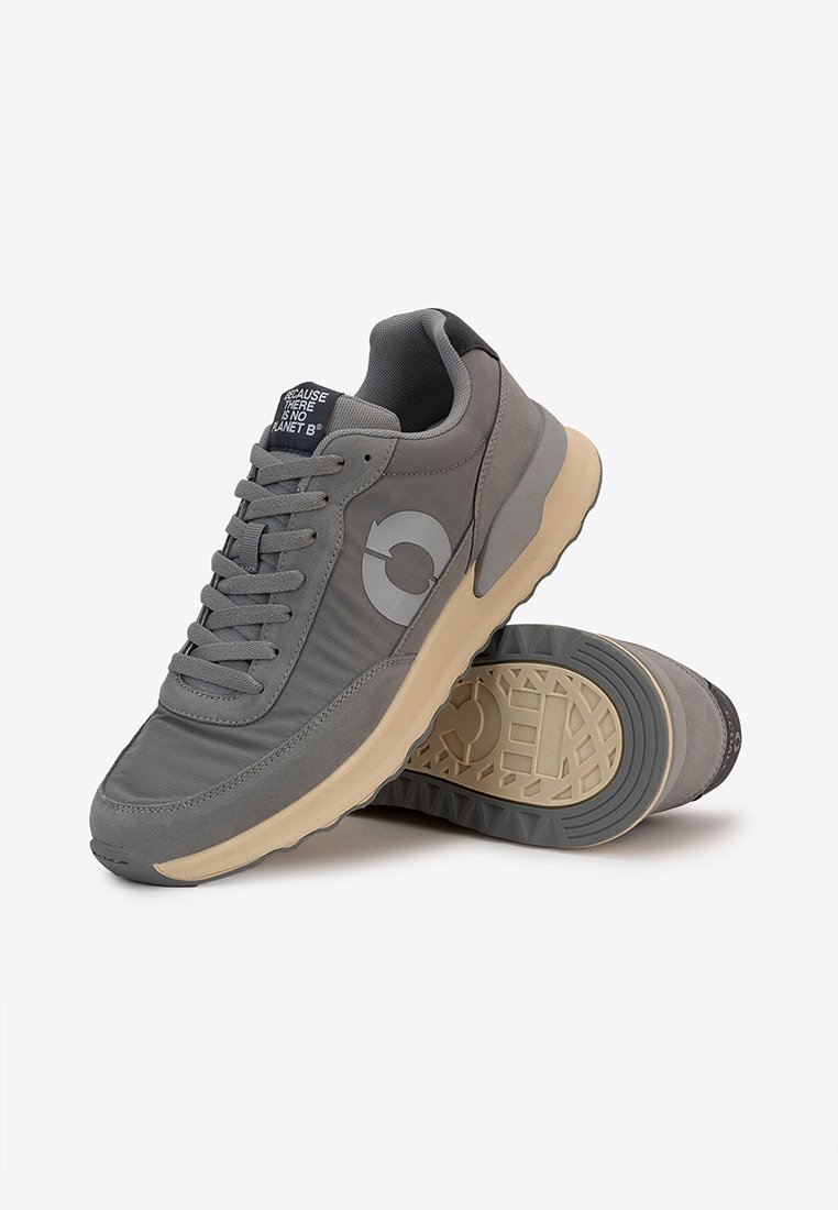 Zapatillas grises con parte superior de tela texturizada, puntera redondeada, diseño con cordones y suela de goma beige en contraste. Presenta un logo en el costado.