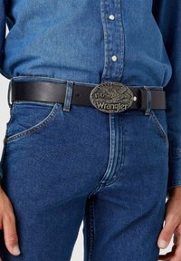 Black leather belt with a large, detailed silver buckle featuring an eagle and the text "1947 Wrangler." Worn with a denim shirt and jeans.