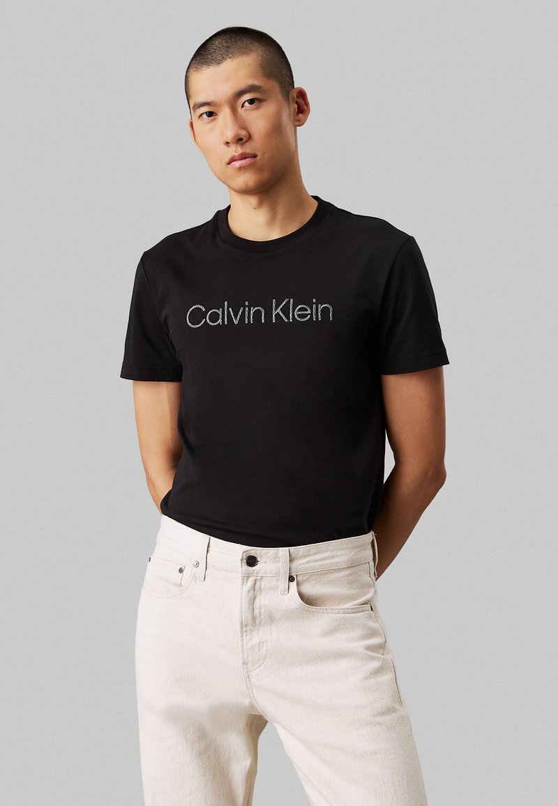 T-shirt en coton noir avec un logo "Calvin Klein" gris au centre, associé à un jean en denim écru, présentant un look décontracté et moderne.
