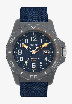Timex Expedition Solar laikrodis turi pilką plastikinį korpusą, mėlyną tekstūruotą audinio dirželį, tamsiai mėlyną bangų rašto ciferblatą ir švytinčias rodykles.