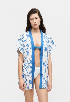 Kobieta ubrana w biało-niebieskie kwiatowe kimono z otwartym przodem, na niebieskim staniku od bikini i białych majtkach od bikini, stoi na jednolitym tle.