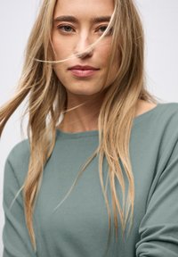 Hellgrüner Pullover mit weicher Textur, weitem Rundhalsausschnitt und langen Ärmeln, ergänzt durch lockeres, welliges, blondes Haar.
