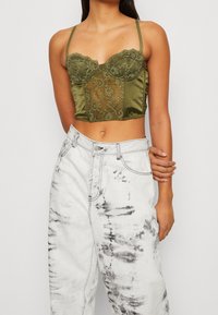 Olivgrön satin- och spetskorsett med justerbara axelband, med en strukturerad byst och detaljerade spetspaneler, åtföljd av vita tie-dye-jeans.