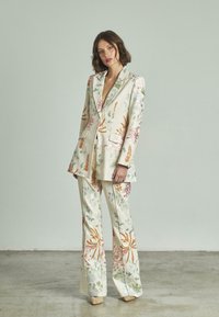 Traje de tela crema con patrón floral; americana ajustada con solapas, combinada con pantalones acampanados. Presenta un bordado colorido en rosa, verde y naranja.