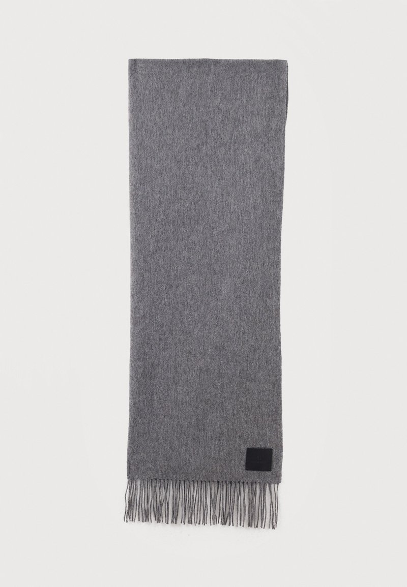 Μαλακό γκρι μάλλινο κασκόλ με φουντωτά άκρα και ένα μικρό μαύρο ορθογώνιο σημείο κοντά σε μία άκρη, στρωμένο επίπεδα σε λευκό φόντο.