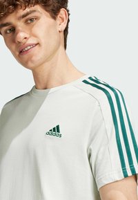 T-shirt bianco in cotone con accenti verdi a tre strisce sulle spalle e logo Adidas ricamato in verde sul petto.