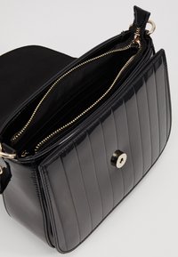 Borsa in pelle verniciata nera con design trapuntato, cerniera dorata e chiusura a scatto magnetica. L'interno è foderato con tessuto liscio.