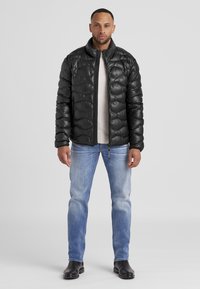 Schwarze Daunenjacke mit gesteppter Optik, hohem Kragen, Frontreißverschluss, über einem hellen Sweater getragen, kombiniert mit hellblauen Jeans und schwarzen Stiefeln.