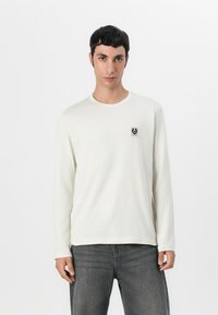 Long-sleeve cream-colored t-shirt, featuring a small black logo patch on the left chest, made of lightweight fabric, with a round neckline.