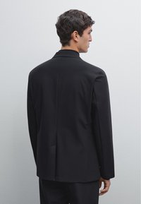 Blazer nero con un design elegante, vestibilità sartoriale, spacco singolo sul retro e texture del tessuto liscia. Non presenta hardware o accenti visibili.