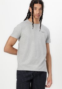 T-shirt gris à manches courtes en tissu doux, avec un petit logo noir sur la poitrine et une coupe décontractée, associé à un pantalon foncé rayé.