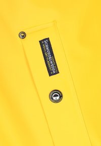 Gul vattentät jacka med en blå tygetikett med text och två cirkulära logotypmärken, ett i svart och ett i vitt.