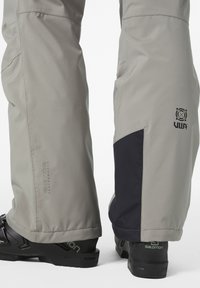 Graue wasserdichte Hose mit schwarzem Einsatz, versehen mit der Helly Tech Marke, getragen mit schwarzen Schuhen, die das Salomon-Logo zeigen.