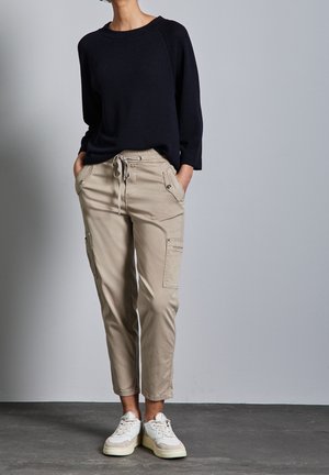 Beige cargobroek met meerdere zakken en een trekkoord in de taille, gecombineerd met een donkerblauwe trui en witte sneakers, tegen een neutrale achtergrond.