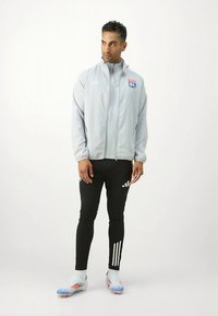 Grijs sportjack met een logo, zwarte strakke broek met witte strepen, en sportschoenen met een blauw-wit ontwerp.