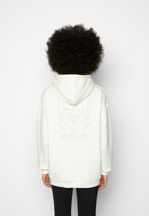 Persoon met krullend haar die een oversized witte hoodie draagt met vage grafische tekst op de achterkant, staand tegen een effen lichte achtergrond.