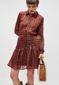 Καφέ φόρεμα με σχέδιο, με γιακά, μακριά μανίκια και φούστα σε διαδοχικά στρώματα. Το ύφασμα έχει γυαλιστερή υφή με μπλε και μαύρες λεπτομέρειες.