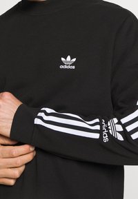 Felpa nera a maniche lunghe con logo adidas bianco sul petto, caratterizzata da tre strisce bianche lungo le maniche. Realizzata in un tessuto liscio.
