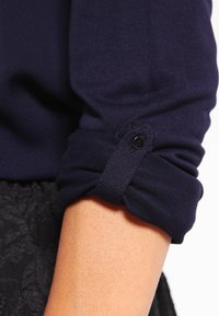 Manche longue bleu foncé retroussée avec une patte de boutonnage noire maintenant le poignet au-dessus d'un avant-bras nu.