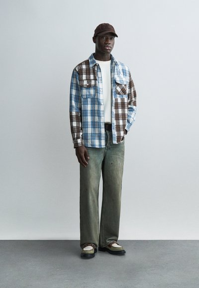 Muž stojící v hnědé a modré kostkované košili, bílém tričku, vybledlých zelených kalhotách, béžovo-černých botách a hnědé čepici na šedém pozadí.