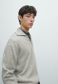 Hellgrauer, gerippter Pullover mit Reißverschluss am Kragen, der eine weiche Textur und eine lockere Passform bietet. Trägt man über einem dunkleren Hemd mit Kragen.