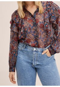 Femme portant un chemisier transparent à fleurs avec des volants et un jean bleu clair taille haute, une main dans la poche, debout devant un fond uni.
