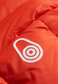 Oranje pufferjack stof met een glanzende textuur, voorzien van een wit logo van concentrische cirkels en een avocado-achtige vorm.