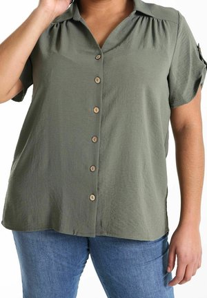 Person wearing an olive green short-sleeve button-up shirt with blue jeans, hand resting near shoulder, cropped at mid-torso.