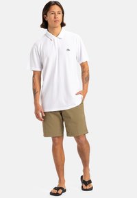 Weißes Polohemd mit Kragen und kurzen Ärmeln, kombiniert mit hellbraunen Shorts. Verfügt über ein kleines schwarzes Logo auf der Brust. Schwarze Flip-Flops.