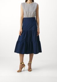 Mujer vestida con una blusa sin mangas blanca y azul marino a rayas, una falda midi azul marino con volantes y sandalias de tacón alto doradas, de pie sobre un fondo blanco.