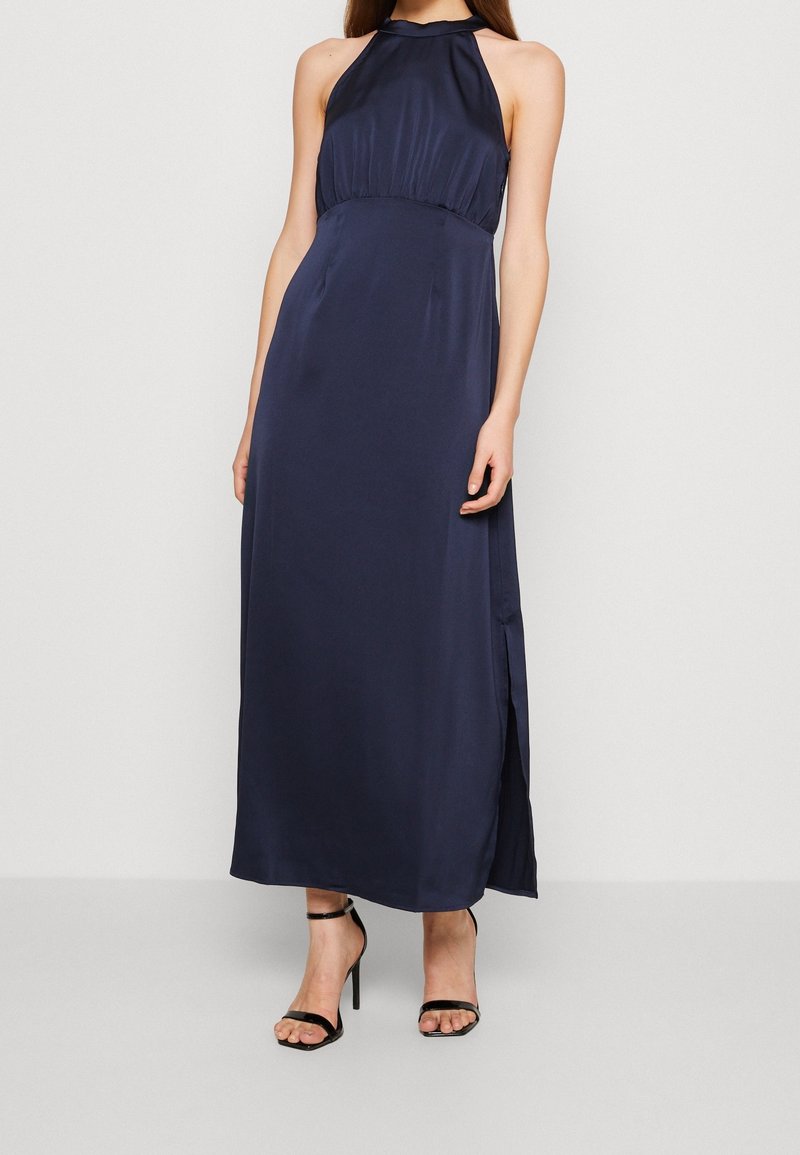 Robe longue sans manches bleu marine en satin, avec un corsage ajusté, une taille froncée et une fente latérale. Associée à des talons noirs à lanières.