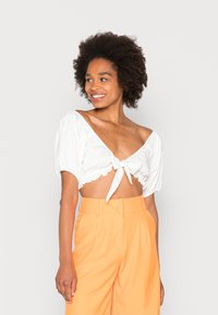 Blusa branca curta com mangas bufantes, detalhe de laço na frente e bainha com bicos. Combinada com calças largas laranja de cintura alta.