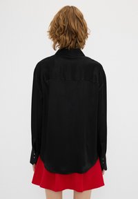 Camicia nera di raso con orlo curvo, maniche lunghe e polsini con bottoni, abbinata a una gonna rossa a campana, vista da dietro.