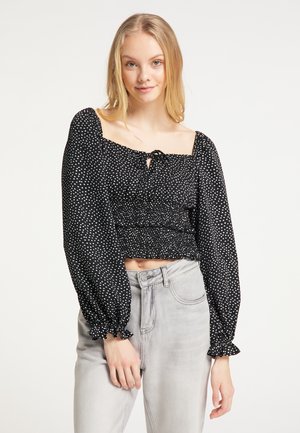 Junge Frau mit langen blonden Haaren, die ein schwarzes, langärmeliges, gerafftes Crop-Top mit weißen Punkten und hellgraue High-Waist-Jeans trägt.