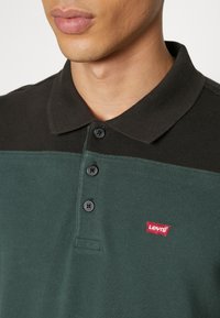 Polo tričko v tmavě zelené a černé barvě, s tříknoflíkovou légou, měkkou texturou a červeným logem Levi's na levé části hrudi.