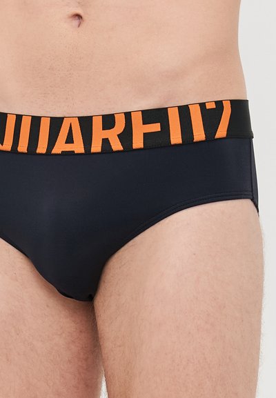 Männlicher Oberkörper, der schwarze Unterwäsche mit einem orangen und schwarzen elastischen Bund trägt, der teilweise fett gedruckte Buchstaben aufweist.