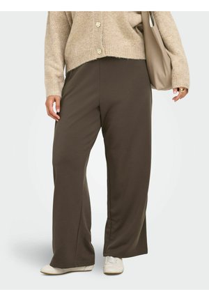 Femme portant un pantalon ample marron foncé, un cardigan beige, des baskets blanches, tenant un sac cabas beige, debout sur fond blanc.